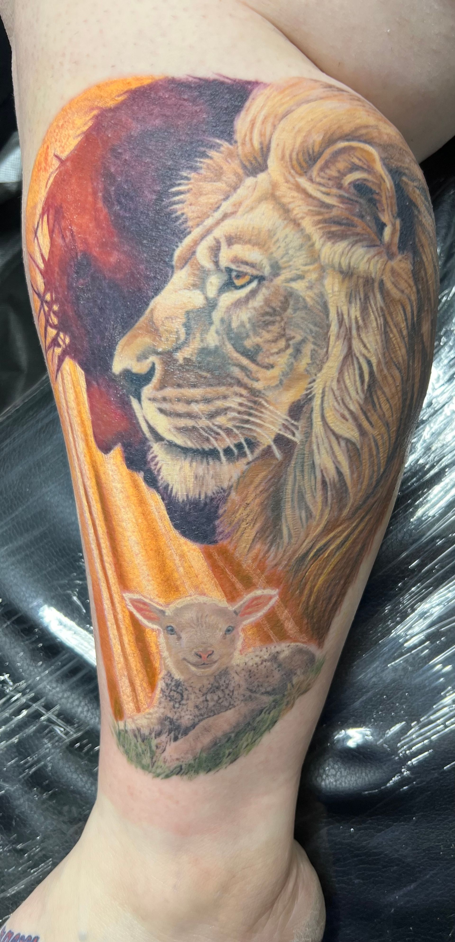Tattoo of a lion and lamb on a leg, with a sunset background.