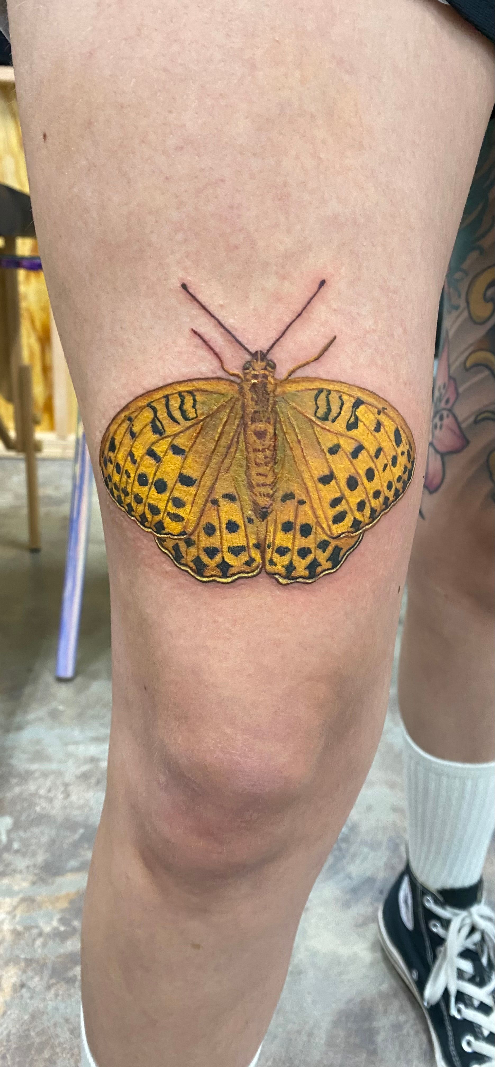 Butterfly tattoo on a person's knee, black and yellow detailed design, with a person wearing black shoes and white socks.