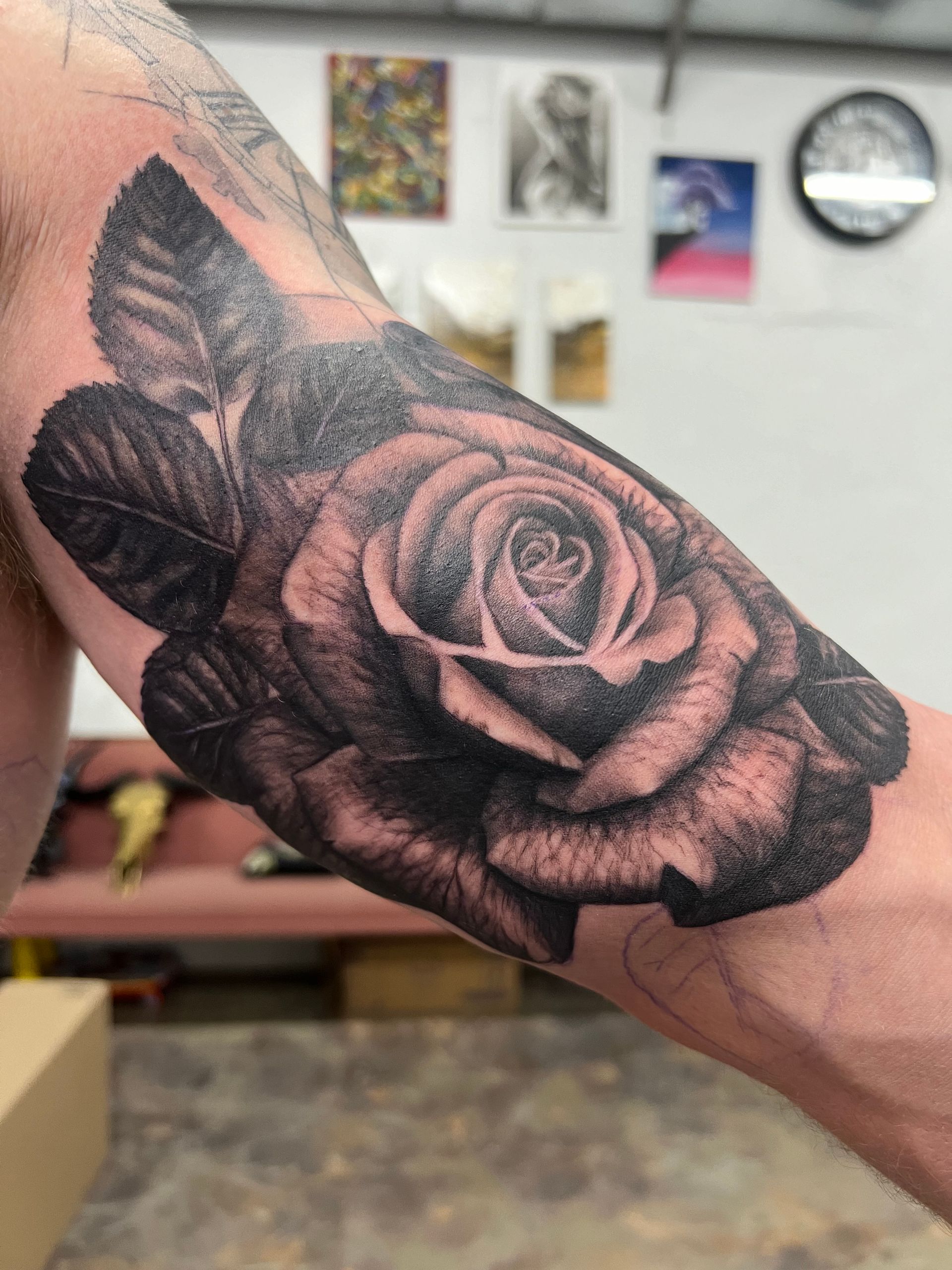 Black and grey rose tattoo on a person's forearm, surrounded by leaves. Located in a shop.