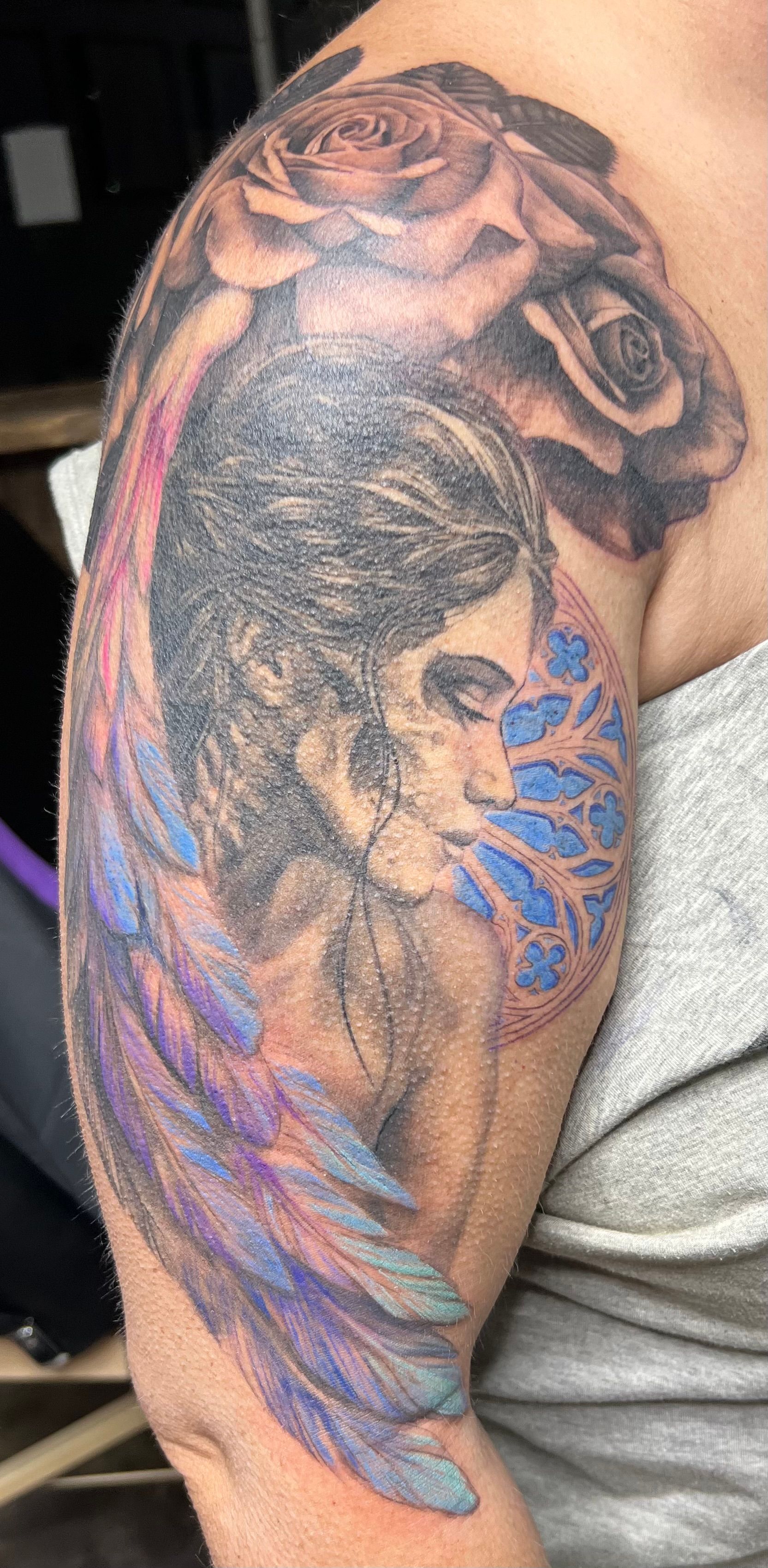 Tattoo of a woman with wings and roses on a person's upper arm; gray, purple, and blue.