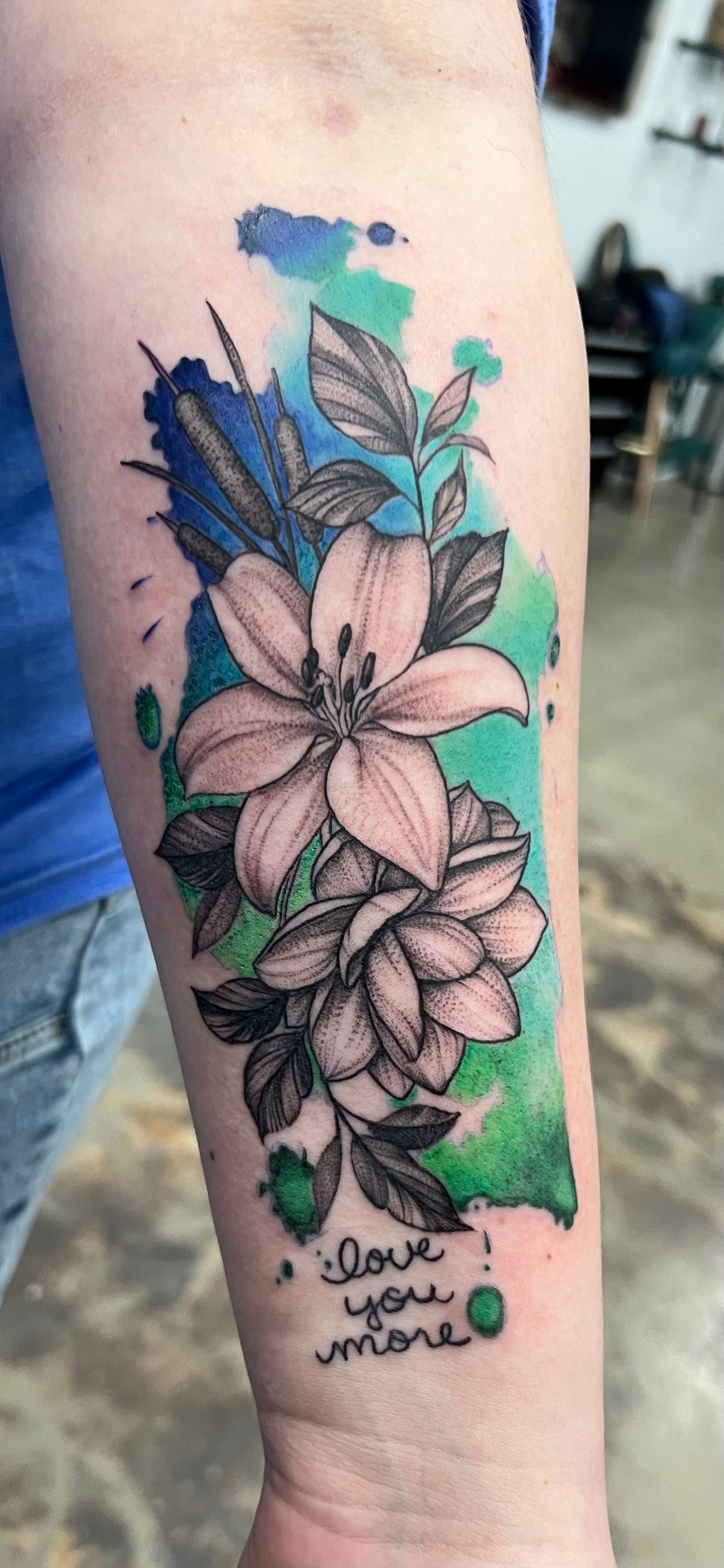 Tattoo of flowers with a watercolor background, blue, green and black ink, text reads