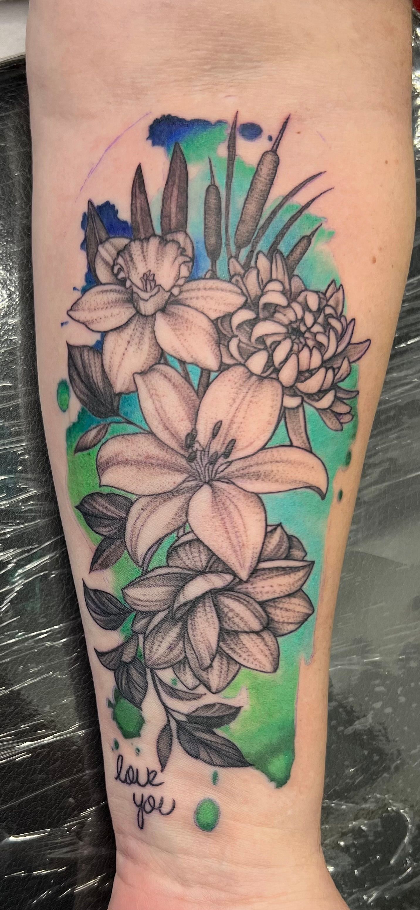 Tattoo of flowers and cattails in black ink with blue and green watercolor. Reads
