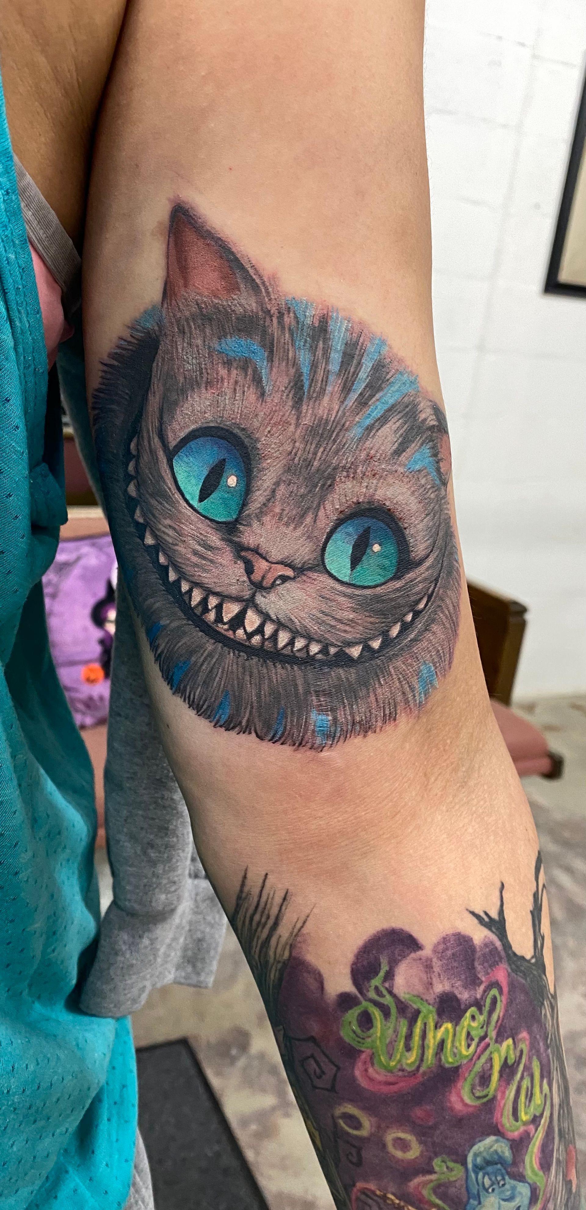 Cheshire Cat tattoo with bright blue eyes on arm.