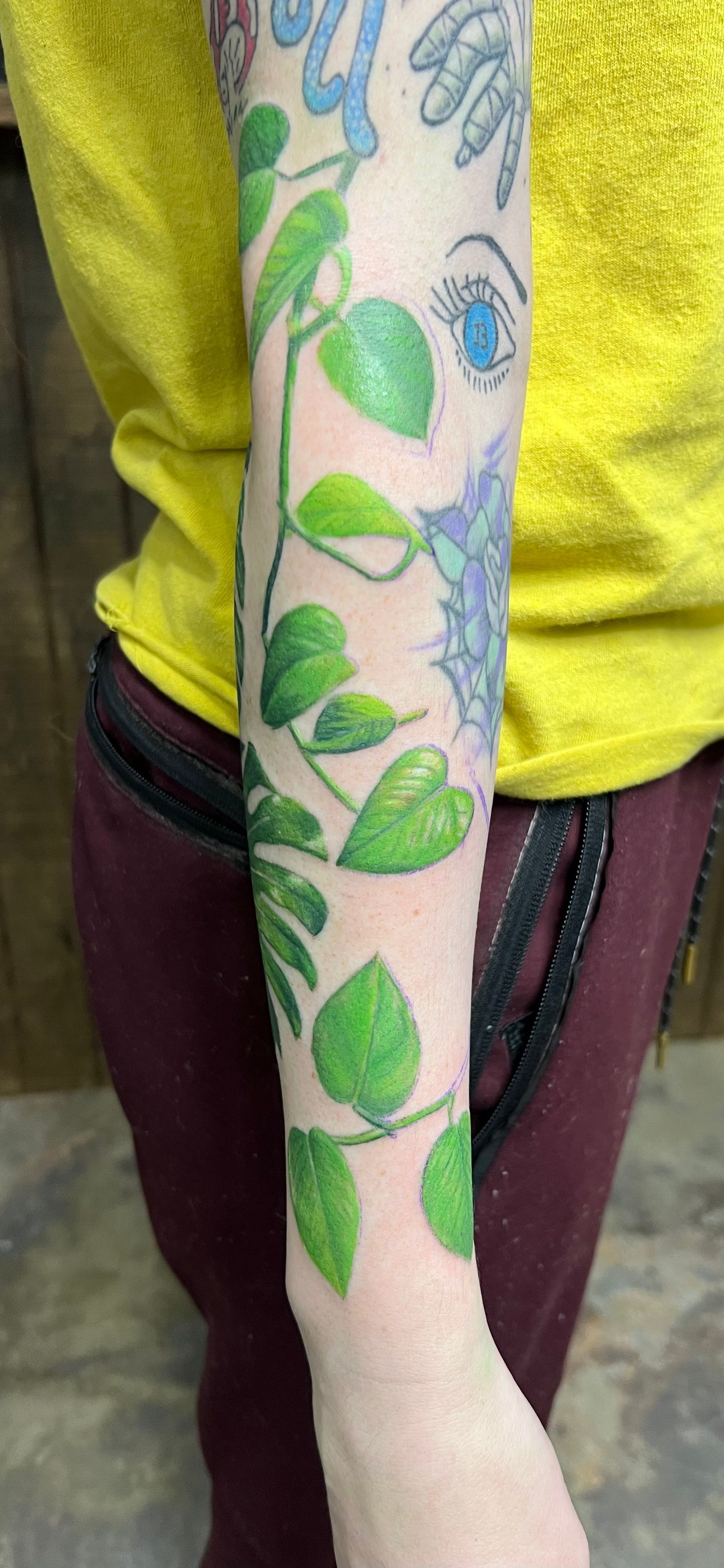 Arm with green vine tattoo, person wearing yellow shirt and burgundy pants.