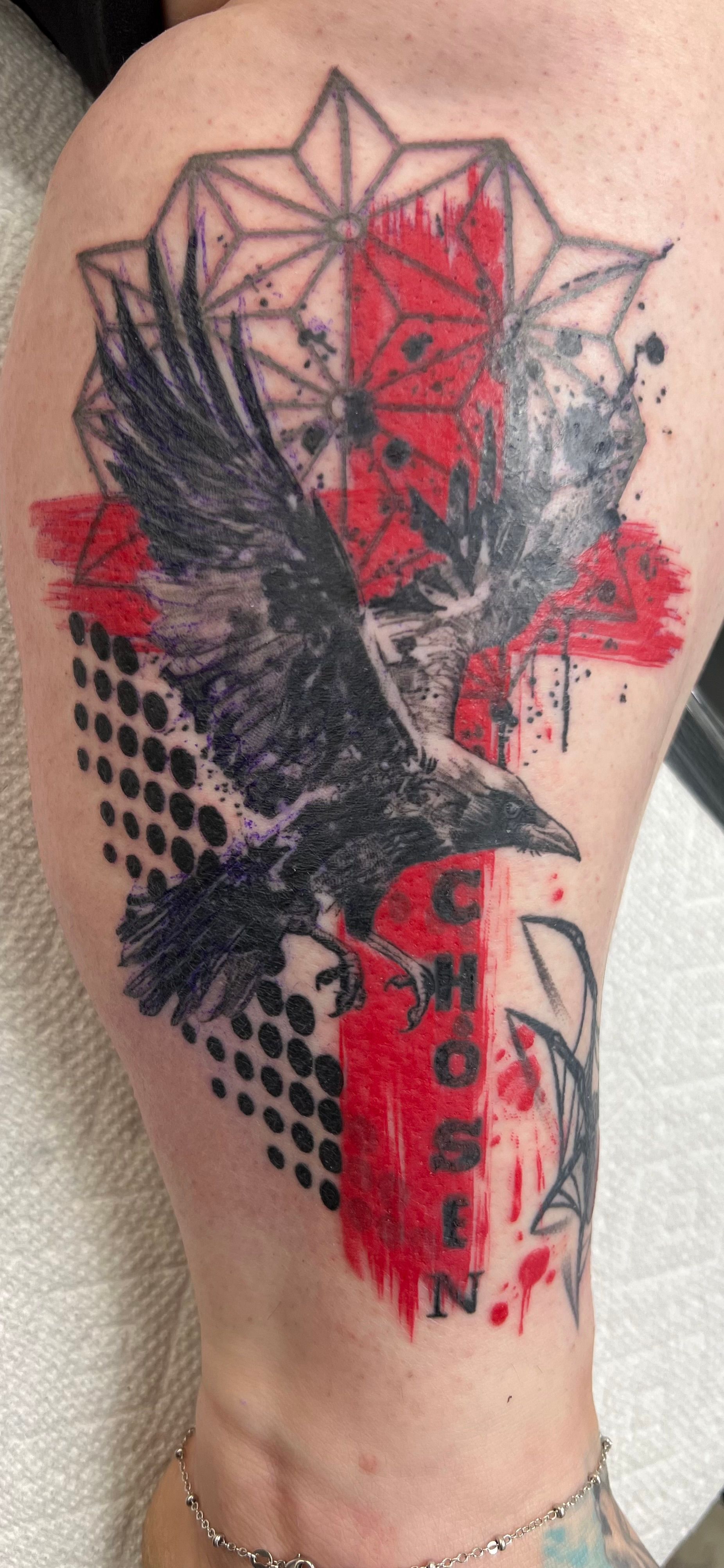Tattoo of a raven with red, black, and white geometric shapes on a leg.