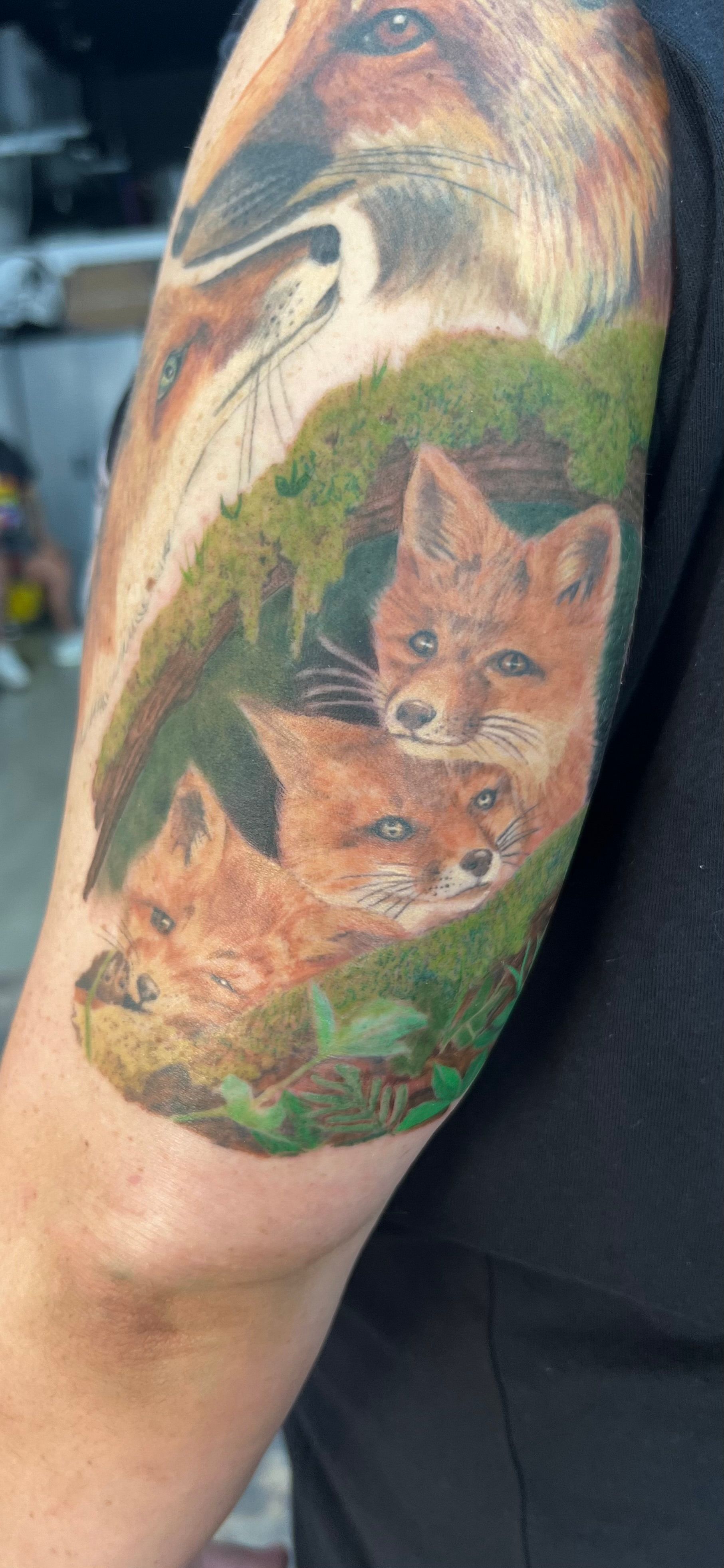 Tattoo of a fox family, with three kits, in a green den on a person's arm.