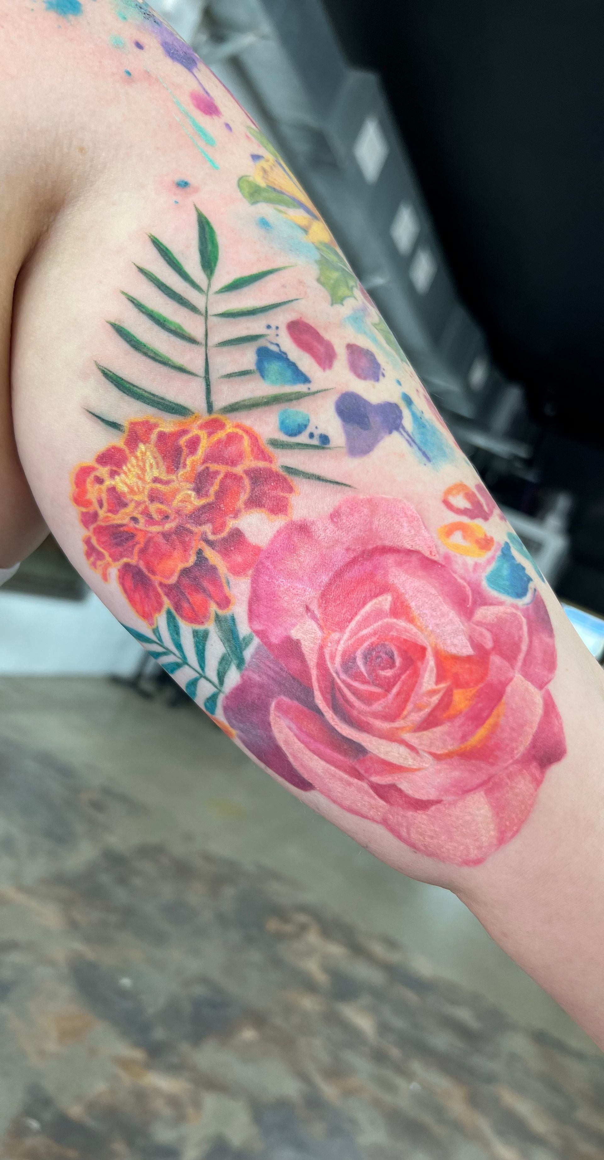Watercolor floral tattoo on an arm: pink rose, orange marigold, fern, blue & purple splashes.