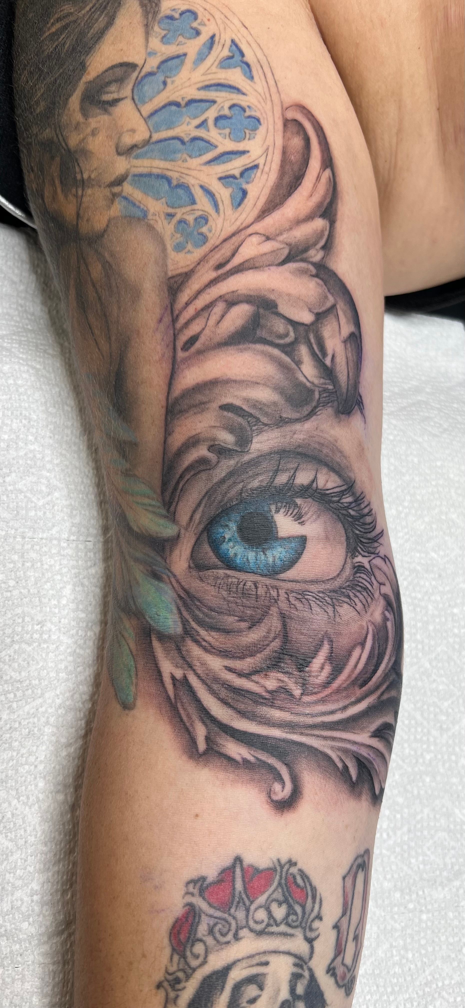 Tattoo of a blue-eyed eye surrounded by ornate details, on a person's arm. A woman's face is above the eye.