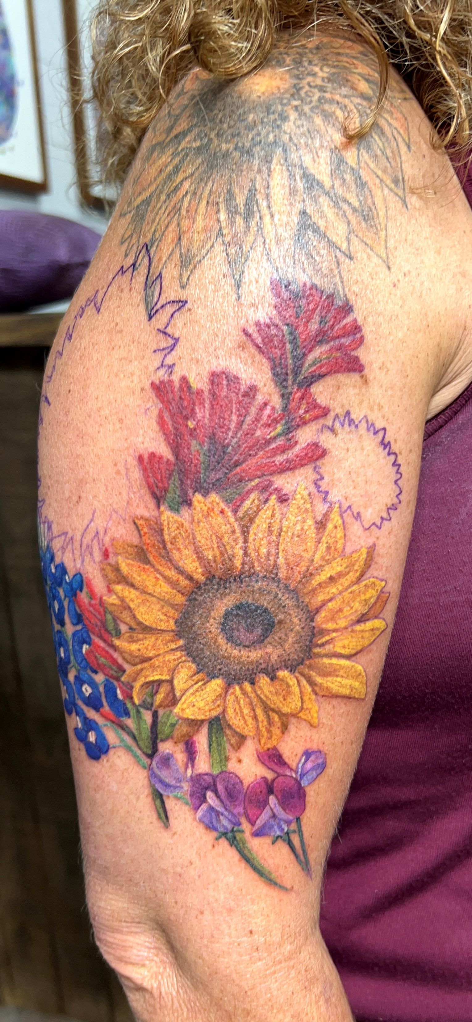 Tattoo of a sunflower, red flowers, bluebonnets, and purple flowers on a person's arm.