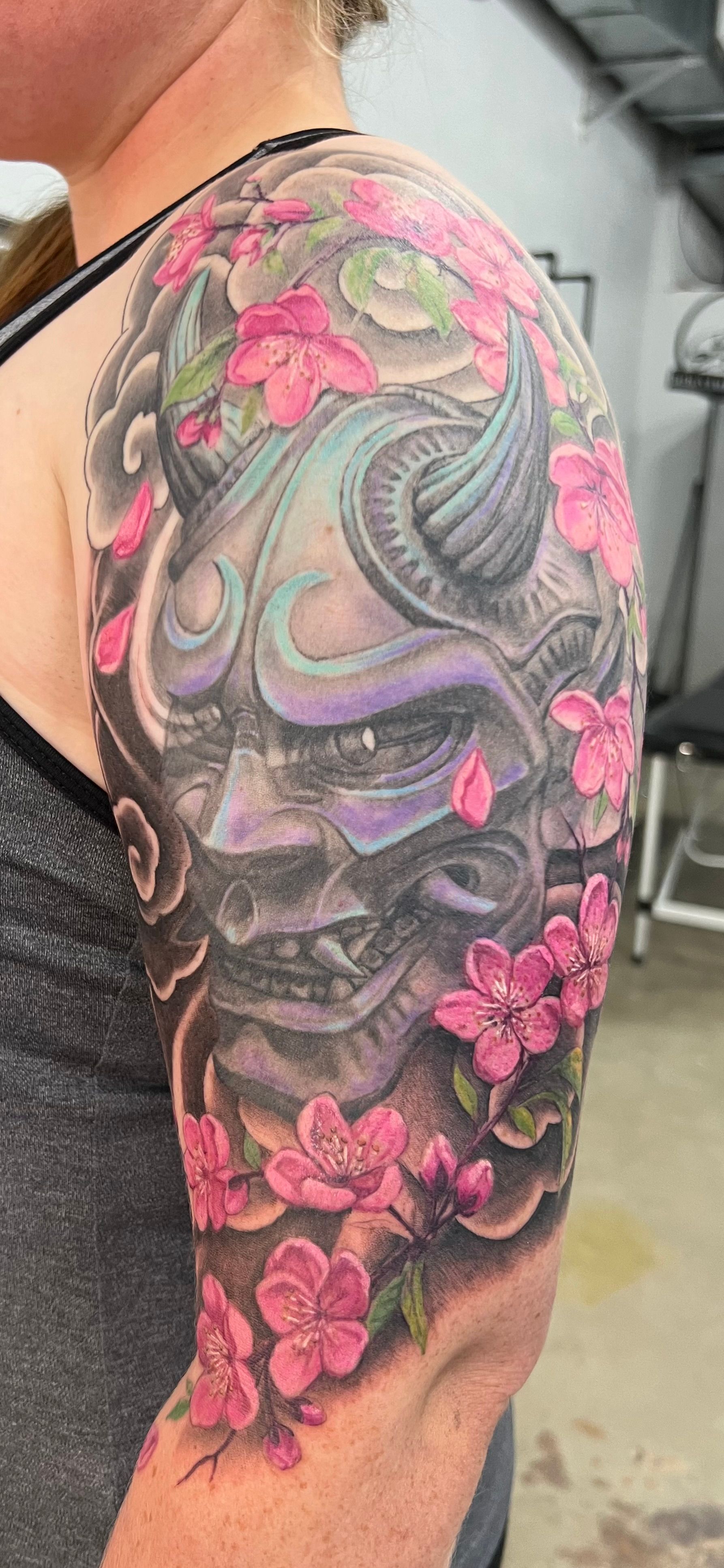 Tattoo of a Japanese demon mask surrounded by pink cherry blossoms on a person's arm.