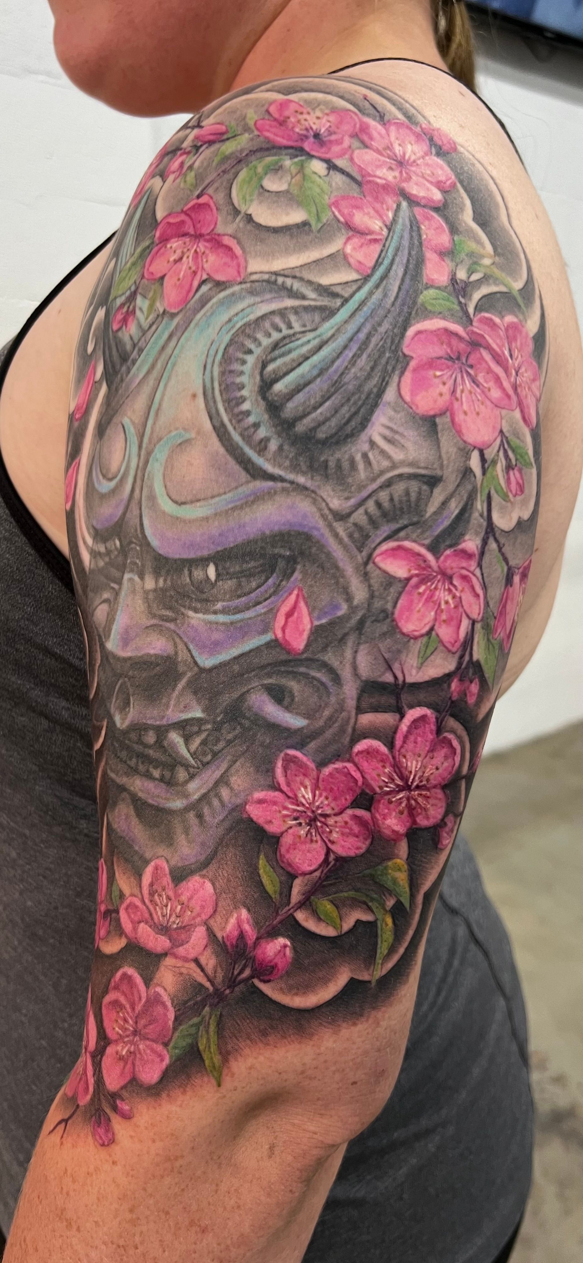 A person's arm tattoo featuring a devil mask with cherry blossoms in a gray, purple, and pink color scheme.