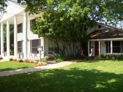 White-columned building with a porch, red door, and trees beside a green lawn