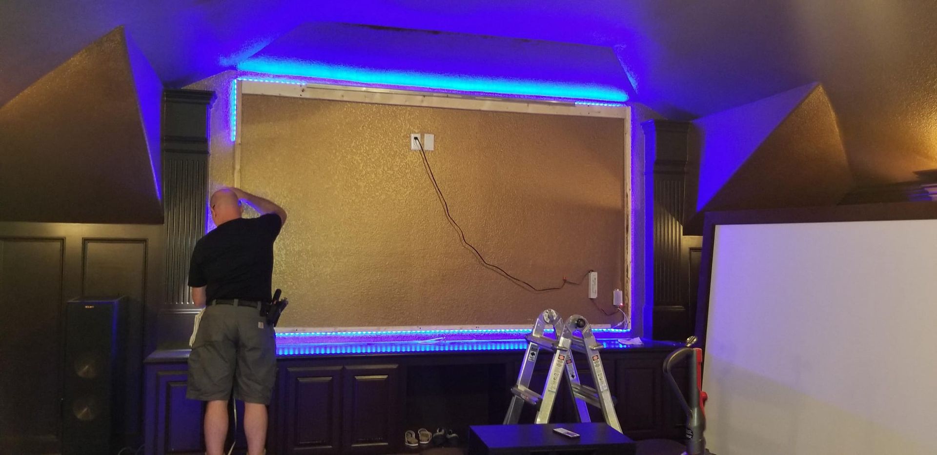 A man adjusts the TV mount frame on a gold wall with blue lights.