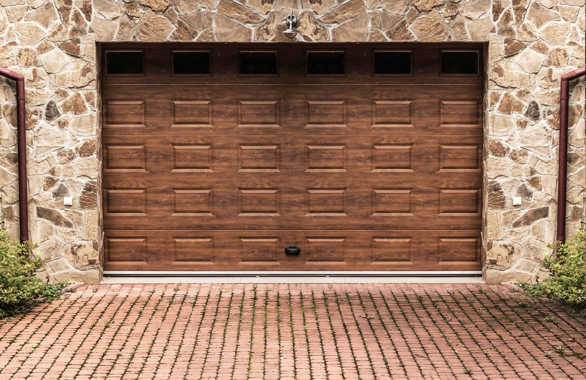 Brown garage door set in a stone wall, on a brick paved driveway.