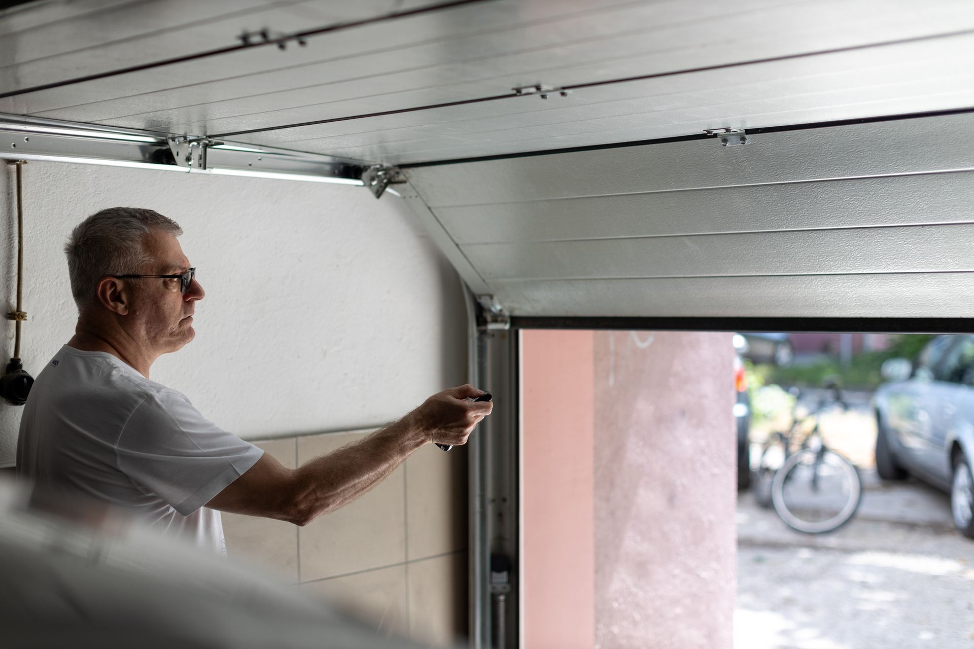 Man in a white shirt operates garage door opener, opening the door. A car and bike are visible outside.