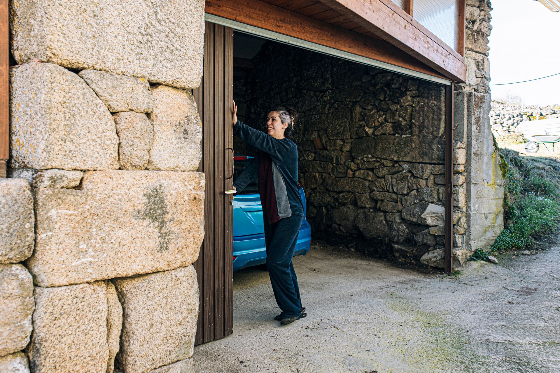 Person in dark clothing stands in a garage doorway, opening the door next to a blue car, stone walls.