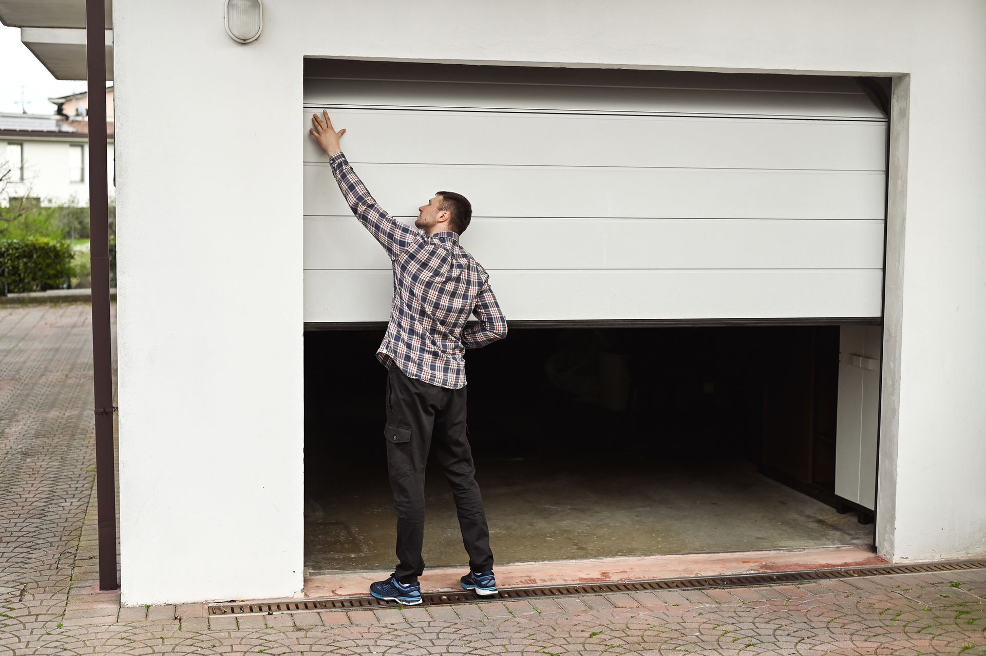 Man opening a white garage door, reaching upward. He's wearing a plaid shirt and dark pants.