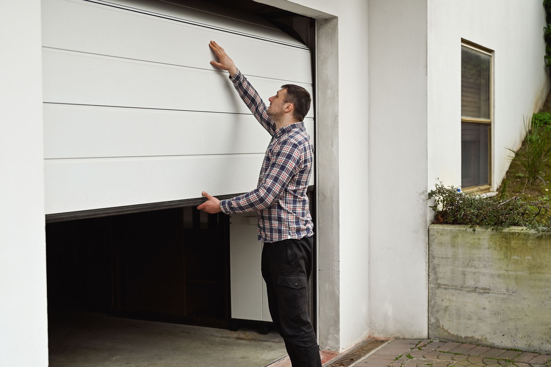 Man inspecting a white garage door, indoors. He's touching the top as it opens, wearing a plaid shirt and dark pants.