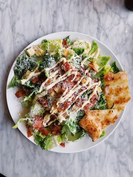 Caesar salad with bacon crumbles, dressing, and grilled bread on a white plate, marble surface.