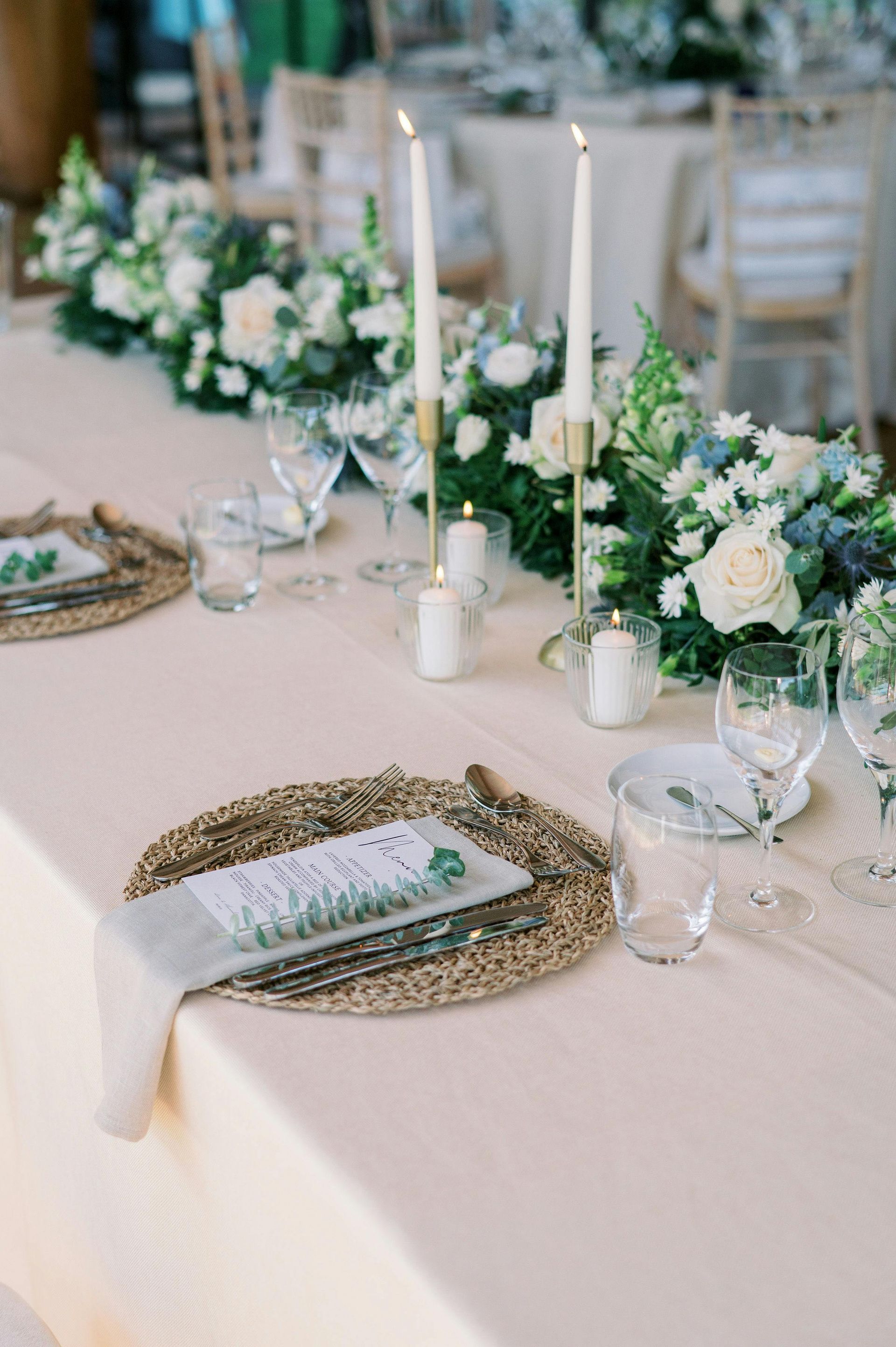 Wedding reception table with floral centerpieces, candles, place settings, and light beige tablecloth.