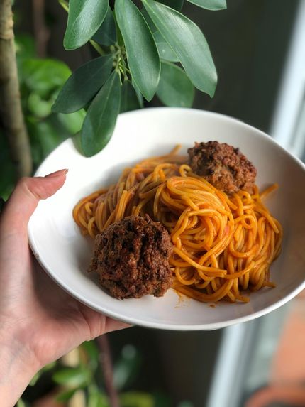 Hand holding a white bowl of pasta with orange sauce and three meatballs, against a green leaf background.