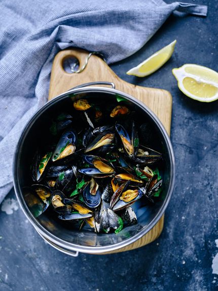 Steamed mussels in a metal pot, wooden board, lemon wedges, and blue cloth.