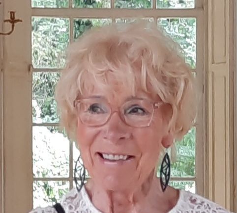 Vaaleat, kiharatukkainen ja silmälasit käyttävä nainen hymyilee ikkunan edessä. Hänellä on korvakorut ja valkoinen toppi.