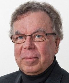 Silmälasit ja musta poolopaita päässä oleva mies katsoo katsojaa yksinkertaista taustaa vasten.