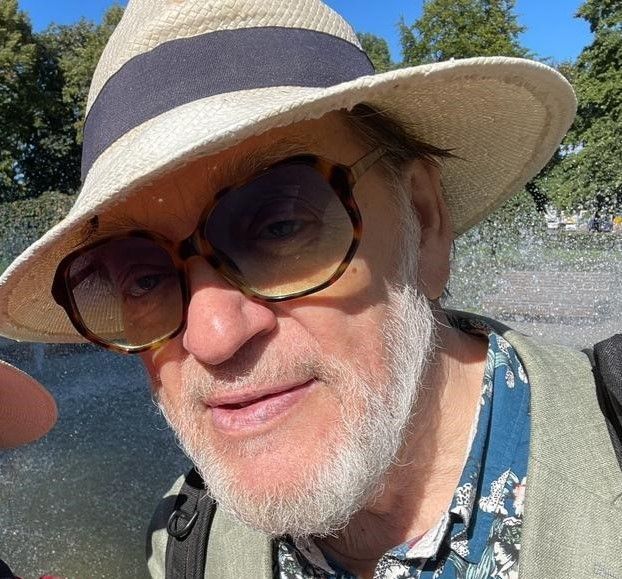 Silmälasit ja hattu päässä oleva mies hymyilee suihkulähteen lähellä.