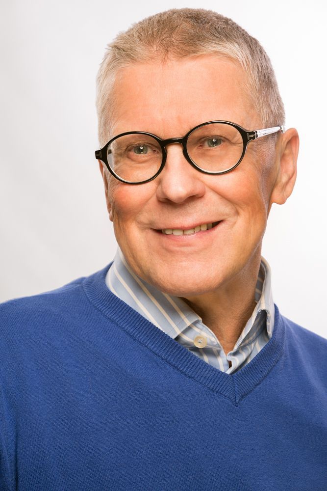 Silmälasipäinen mies hymyilee, yllään sininen neule raidallisen paidan päällä, valkoista taustaa vasten.