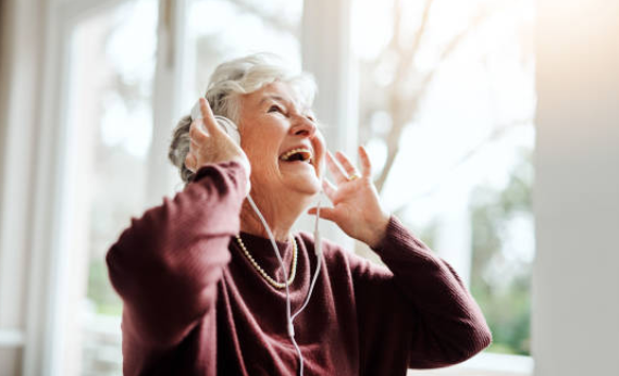 Nainen kuulokkeet päässä, hymyilevä silmät suljettuina, kuuntelee musiikkia. Sisällä, ikkunan lähellä.