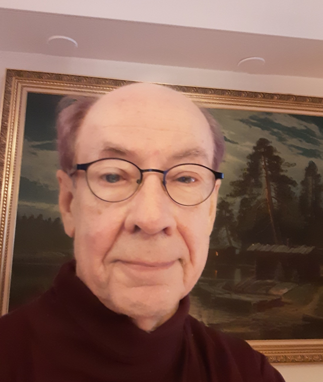 Silmälasipäinen ja punertavanvioletti poolopaidallinen mies kehystetyn maalauksen edessä.