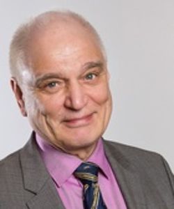 Ohenevat valkotukkaiset hymyilevä mies, jolla on yllään harmaa puvuntakki vaaleanpunaisen paidan ja kuviollisen solmion päällä.