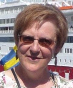 Aurinkolasit päässä oleva nainen seisoo risteilyaluksen edessä, jonka edessä liehuu sinikeltainen lippu.