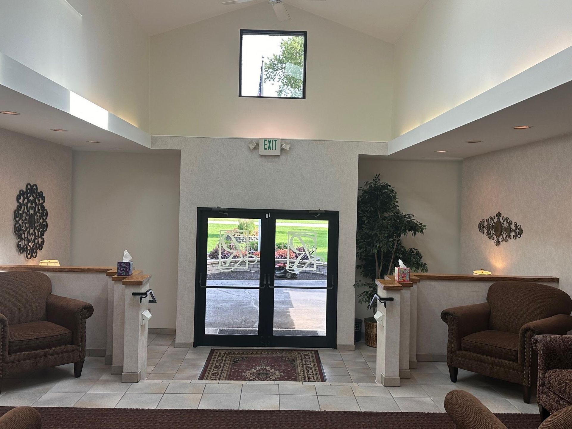 Interior lobby with double glass doors, two armchairs, decorative wall art, and a small window.
