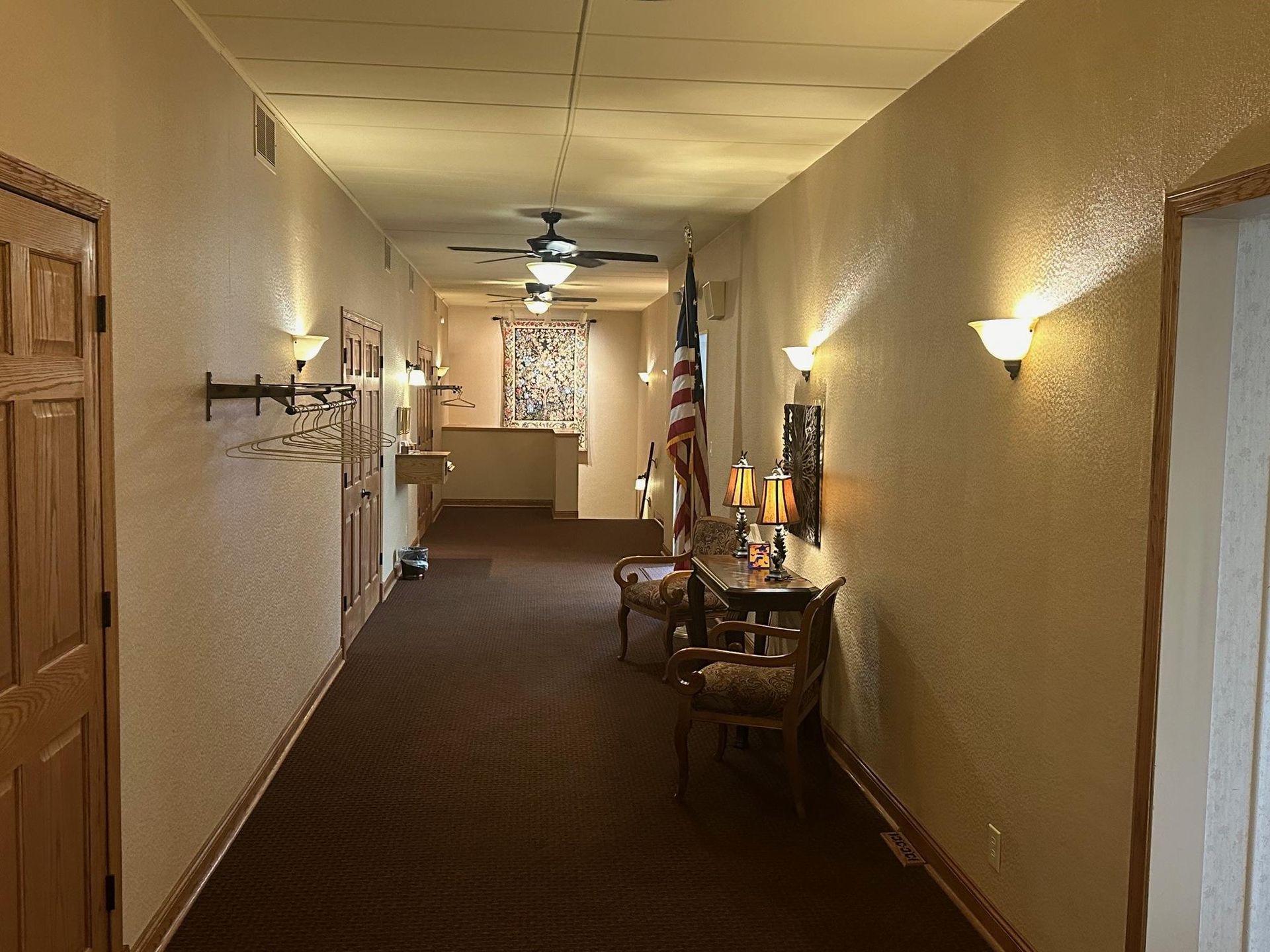 Long hallway with dark carpet, cream walls, sconce lighting, and decorative furniture. American flag on display.