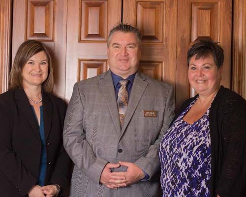 Three people standing in front of a wooden door. The man in a suit, two women with jackets, all smiling.