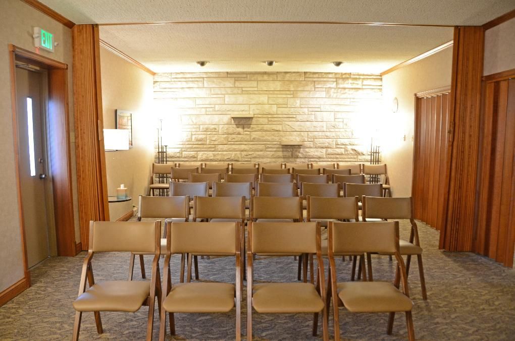 Rows of tan chairs face a stone wall in a dimly lit room with wood trim and curtains.