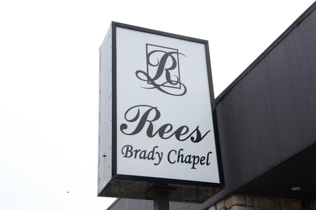 Sign for Rees Brady Chapel; white background, black lettering and logo, attached to building.