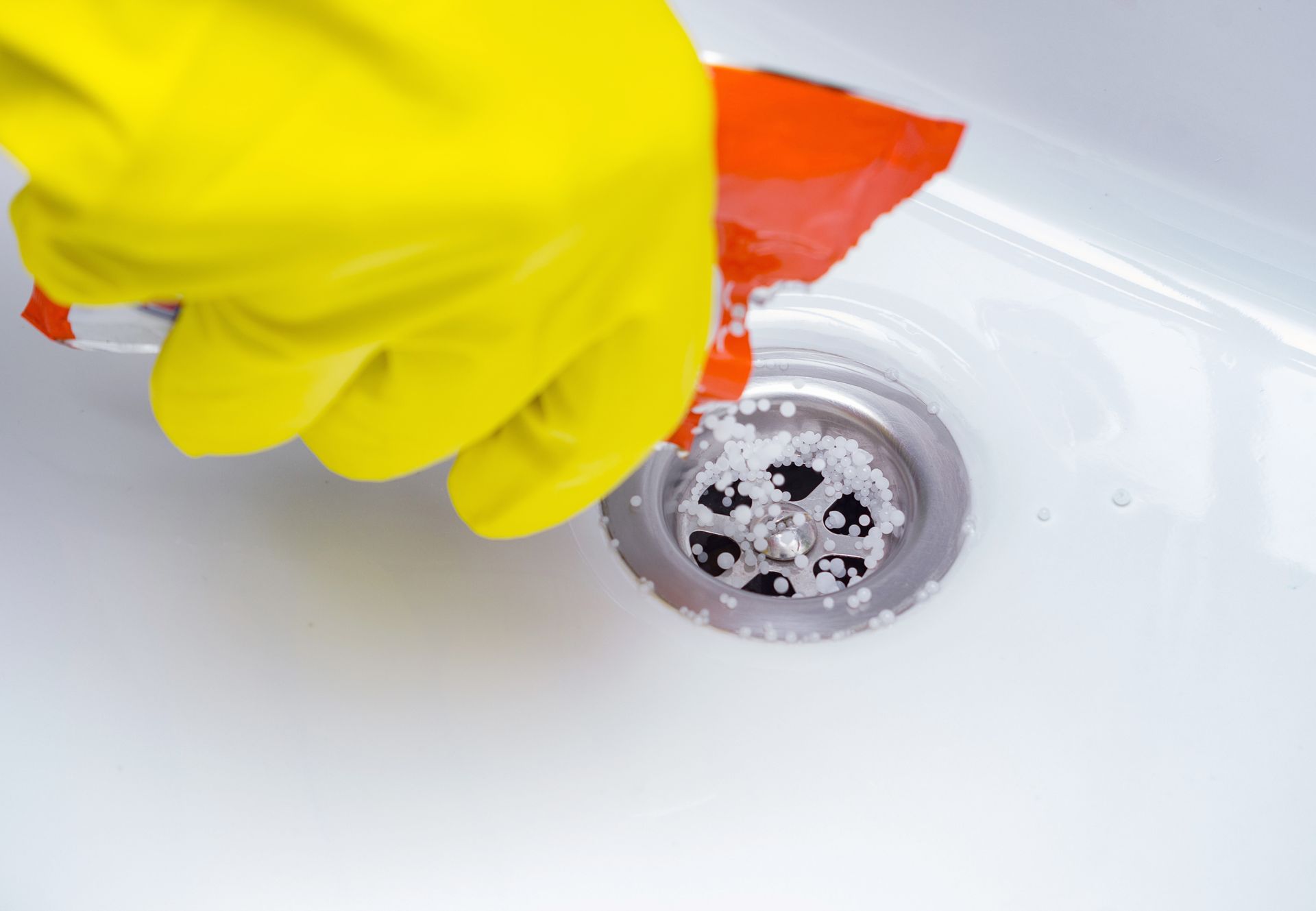 Gloved hand pouring granules into a white sink drain; red and yellow colors.