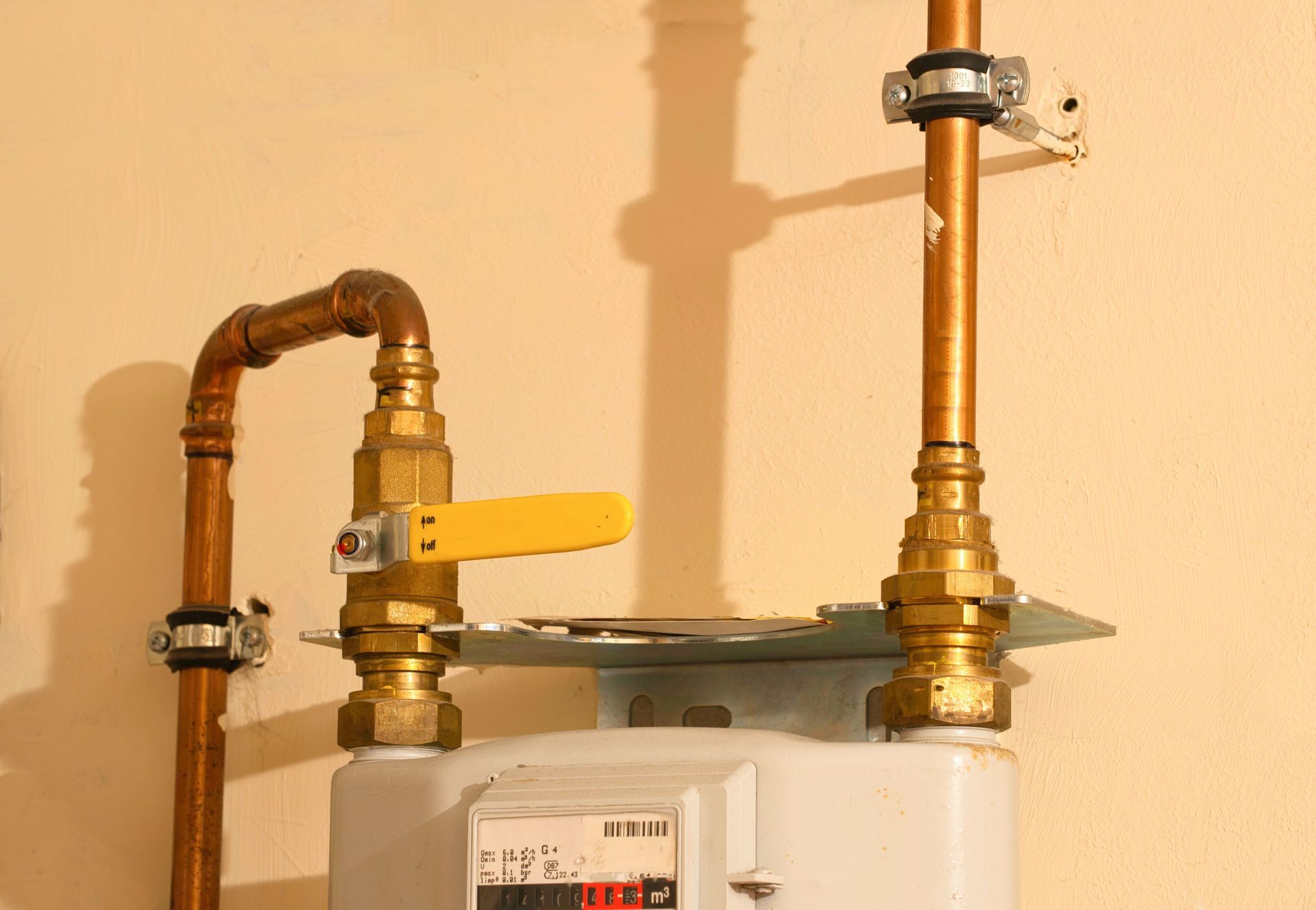 Gas meter with copper pipes and a yellow valve handle against a beige wall.