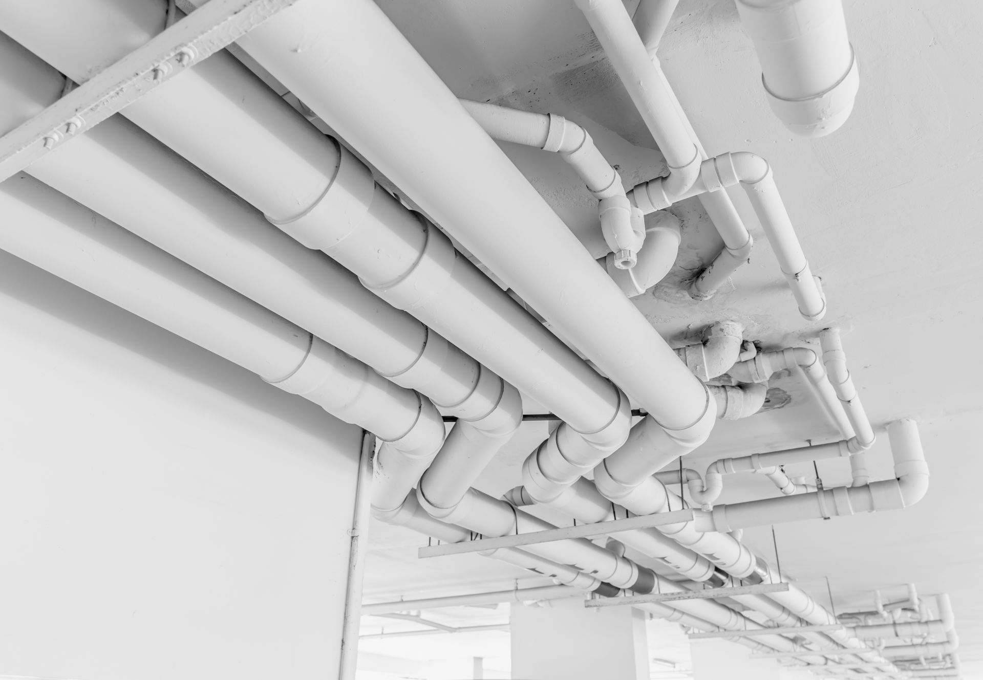 White pipes and fittings on a ceiling, likely for plumbing or utilities, against a white wall.