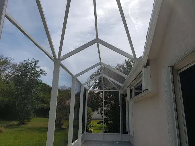 White-framed screened porch with a glass ceiling, attached to a light-colored house, overlooking a green yard under a partly cloudy sky.