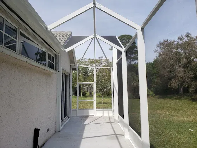 White screened-in patio, attached to a house with a concrete floor, overlooks a green lawn and trees.