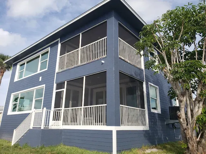 Blue two-story house with white railings and screened balconies. Tree on right, clear sky.