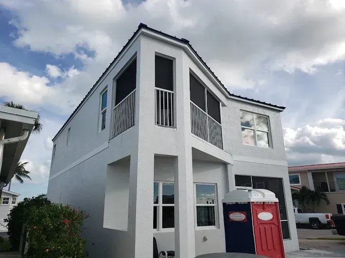 Two-story white building with a dark roof and screened balconies, under a cloudy sky. Two portable toilets in front.