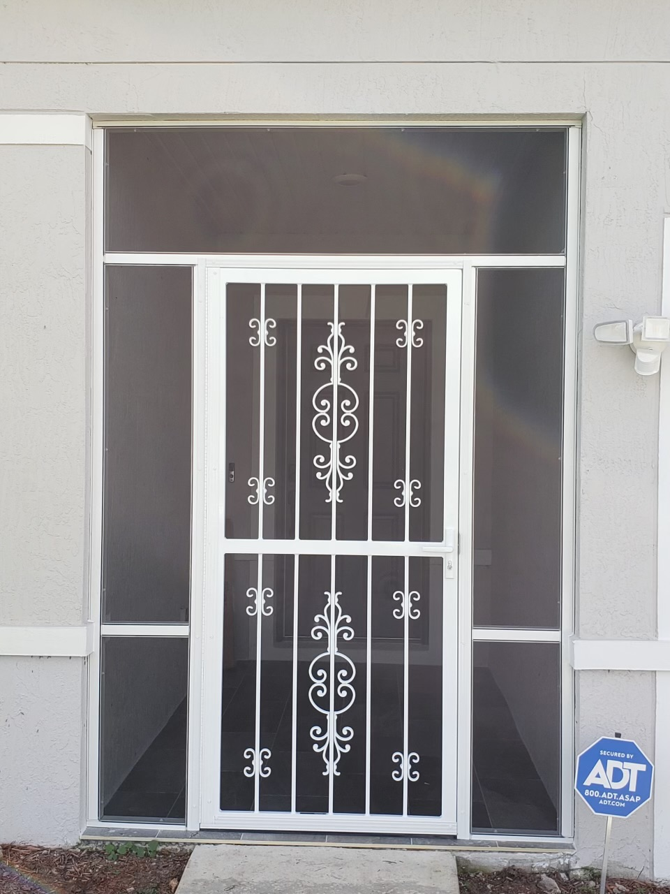 White security door with ornate scrollwork, framed by dark-tinted glass panels, on a gray building.
