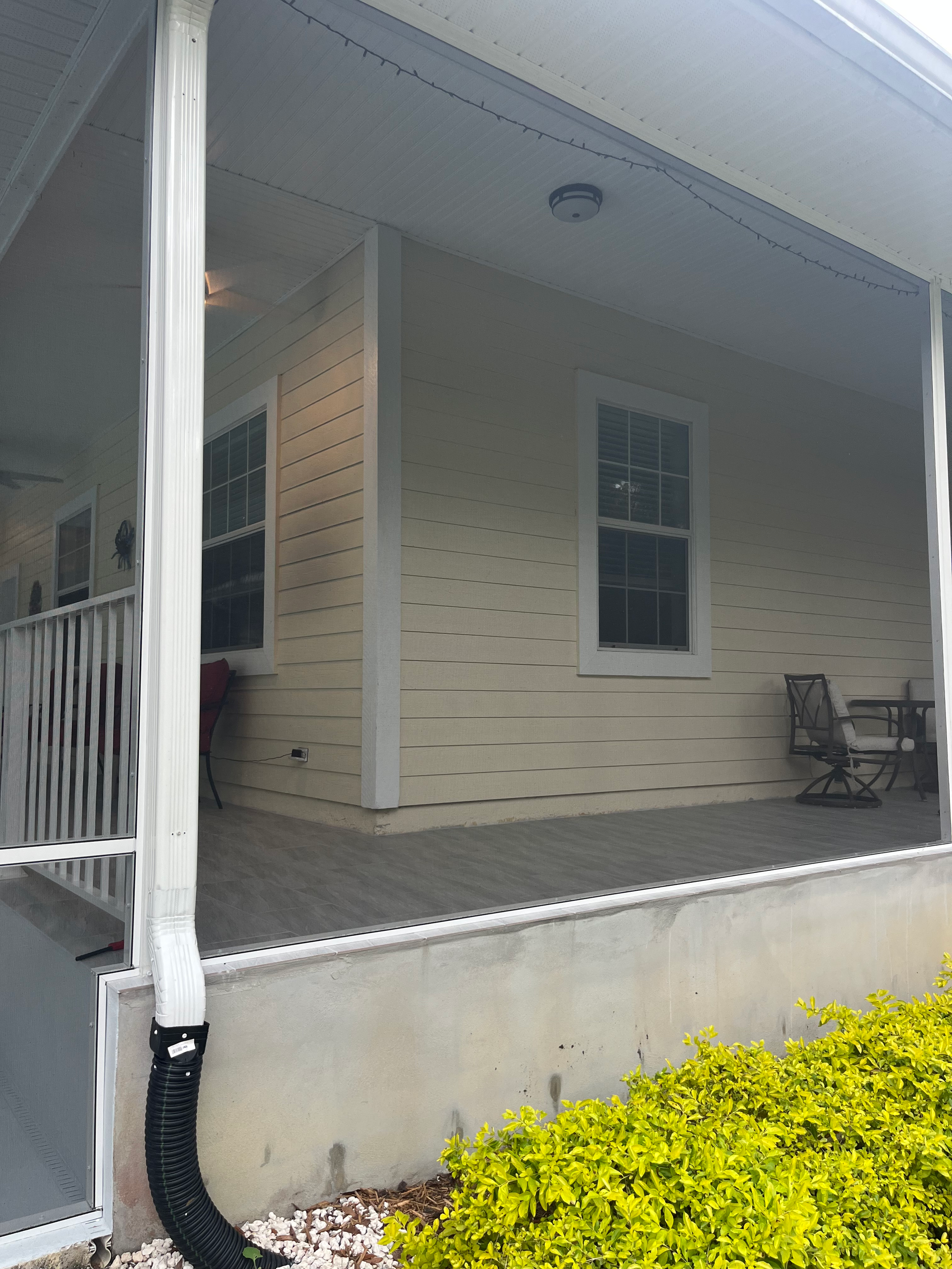 Covered porch with siding and windows, neutral colors, small bushes in foreground.