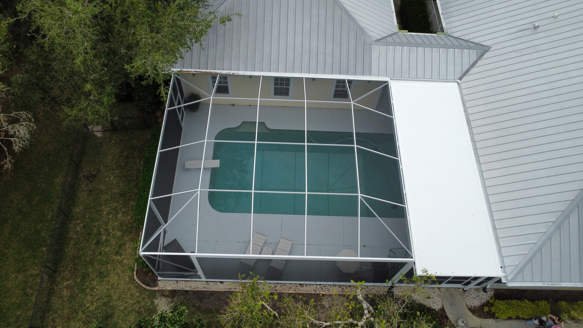 A screened-in patio with white metal frame, set in a yard with green grass and foliage under a partly cloudy sky.