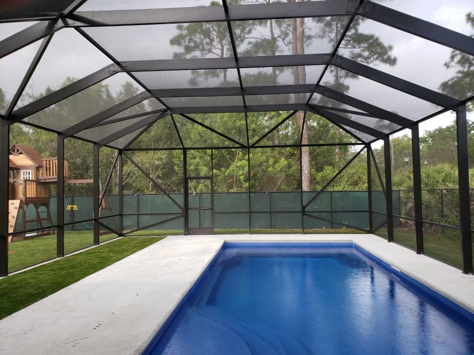 Pool enclosed by a black screened structure. Blue pool, white concrete deck, green grass, and trees.